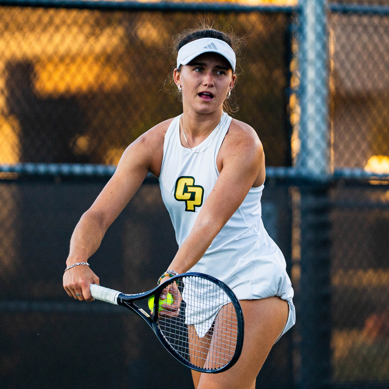A Cal Poly women's tennis team athlete in a white uniform with a CP prepares to serve the ball